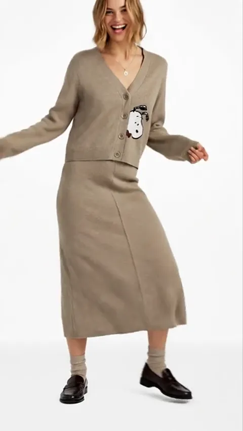 A white studio: a woman in a beige cardigan with an appliqué and a skirt in the same knit, in motion. The set reads as o