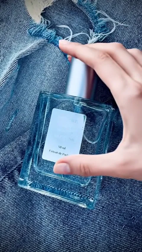 A bottle lies on denim: clear glass, a pale label, a metal cap. A hand sets it down on worn indigo.