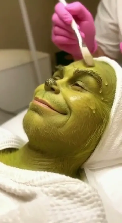 A beautician's room: a green character from a Christmas tale lies on the couch while pink-gloved hands apply a mask with