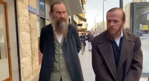 A street in modern Jerusalem: two bearded men in nineteenth-century frock coats walk past shopfronts with Hebrew signs.