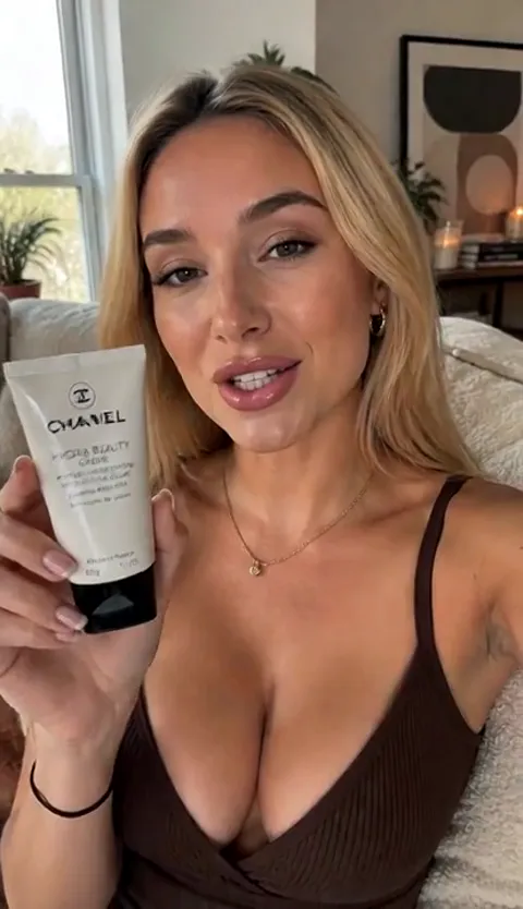 A first-person, blog-style frame: a woman holds a tube of cream to the camera, a bright living room with a window behind