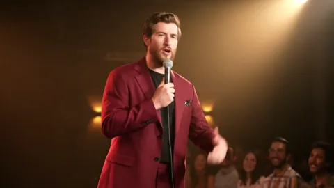 A comedy club stage: a man in a burgundy oversized suit stands with a microphone, warm light and an audience in half-dar
