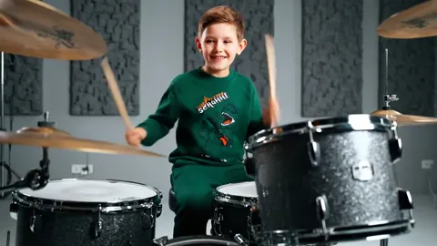 A studio with acoustic panels: a boy in an emerald sweatshirt sits at a drum kit, sticks raised. The sweatshirt is the o