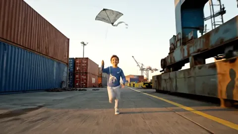 A container port at sunset: a boy in a sweatshirt runs across the concrete toward the camera, a paper plane in his hand.