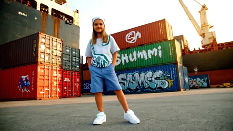 A container yard with SHUMMI graffiti on the sides: a girl in a grey marl t-shirt and denim shorts stands facing us. A d