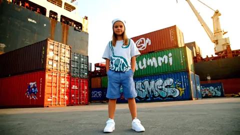 The finale: she stands with the row of containers behind her. Eight seconds have shown the garment from two sides in two