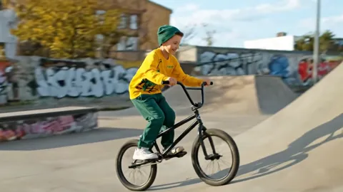 A graffitied skatepark: a boy in a yellow hoodie and green trousers rides a BMX through the bowl. The clothing is the on