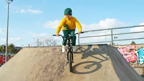 Riding up the ramp, framed from below. The trouser length against the sneakers reads, and the cuff is gathered so it can